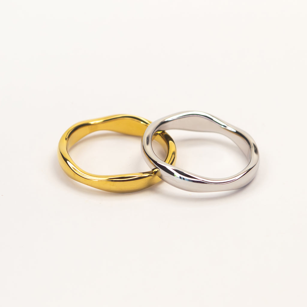 Two wavy, fluid-shaped rings, one in polished yellow gold and the other in polished silver, interlocking on a clean white background.