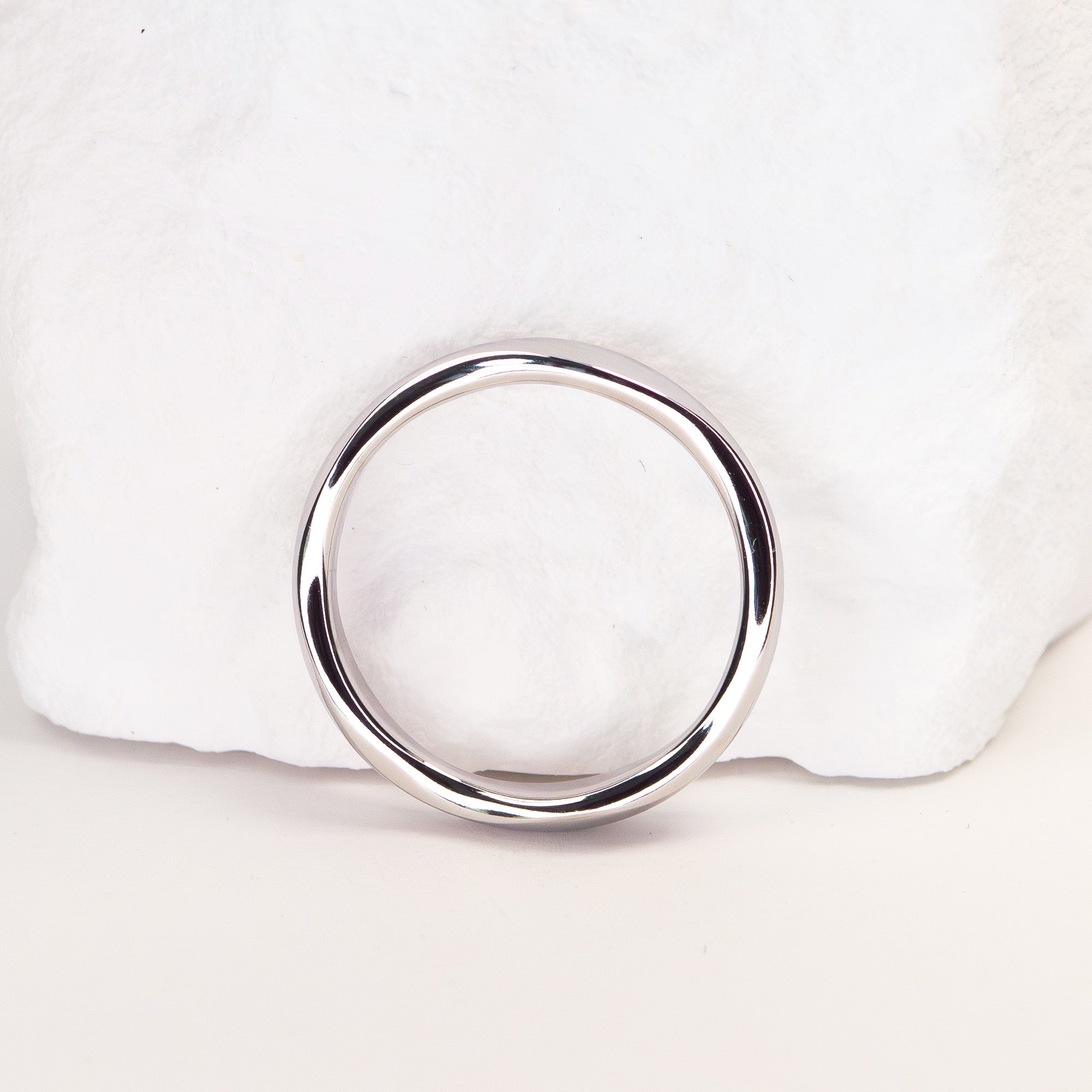 A polished silver-toned metal ring with a thick, rounded band, displayed on a white textured stone prop against a light background.
